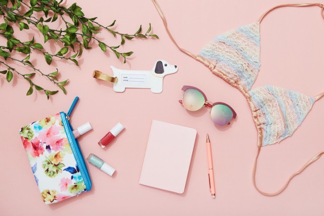 Travel flat lay on pink background with note pad, sunglasses, bikini top, nail polish and luggage tag